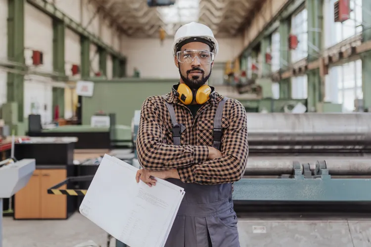 A manufacturing worker stands in a facility with a hardhat and blueprint.